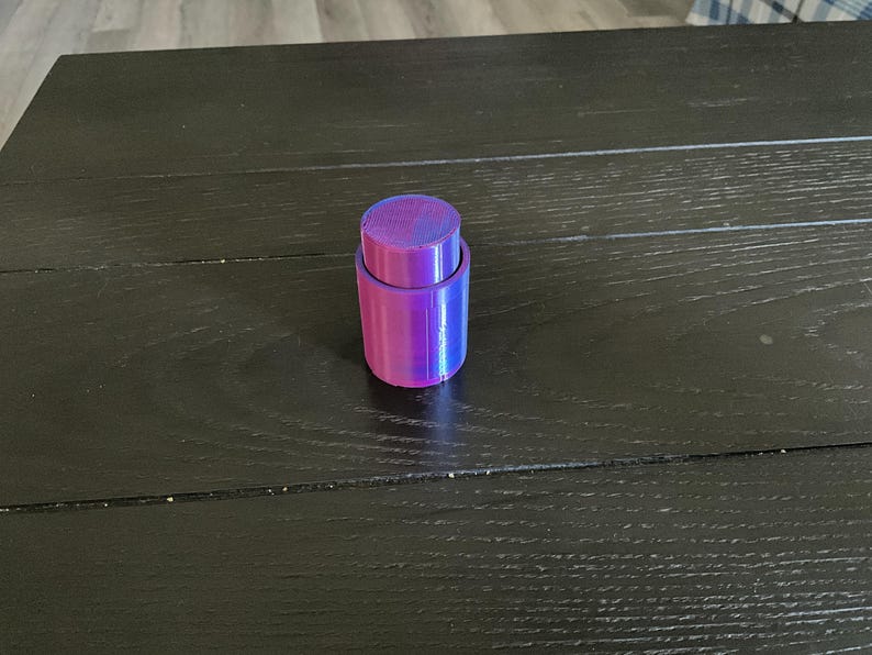 Puede incluir: Un tap&oacute;n de botella iridiscente morado y azul. El tap&oacute;n est&aacute; hecho de dos piezas, una superior y una inferior, que encajan entre s&iacute;.