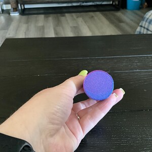Puede incluir: Una esponja de maquillaje redonda, de color morado y rosa, con una superficie texturizada.