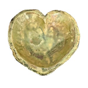 Puede incluir: Cuenco decorativo en forma de corazón con superficie texturizada. El cuenco tiene una combinación de colores verde claro y dorado con un acabado ligeramente iridiscente. Los bordes tienen un aspecto más oscuro y texturizado.