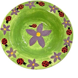Może przedstawiać: Okrągła, ręcznie malowana ceramiczna miska z jasnozielonym wnętrzem. Obrzeże zdobią fioletowe kwiaty i czerwone biedronki z czarnymi kropkami. Duży fioletowy kwiat z żółtym środkiem znajduje się na środku miski.