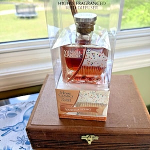 Könnte beinhalten: Ein Yankee Candle Vanilla Icing Reed Diffusor in einer transparenten Kunststoffbox. Die Glasflasche hat ein Cupcake-Design. Die Box steht auf einer Holzbox mit Messingverschluss. Der Text "YANKEE CANDLE" und "VANILLA ICING" sind sichtbar.