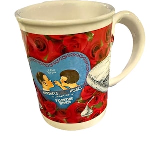 May include: White ceramic mug with a red rose pattern and a blue heart-shaped graphic featuring a couple and the text "Hershey's Kisses Valentine Wishes". The mug has a white handle and rim.