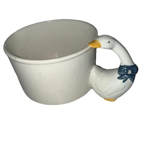 May include: White ceramic mug with a goose-shaped handle. The handle features a goose with a yellow beak and a blue bow. The mug has a slightly flared shape and is suitable for hot or cold beverages.