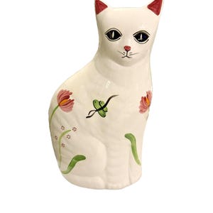 May include: A white ceramic cat ornament with red-tipped ears, black eyes, and a pink nose. The cat is decorated with pink and green floral accents. The ornament is a decorative item.