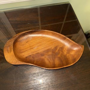 Puede incluir: Una fuente de madera con forma de hoja, de color marrón intenso y con vetas de madera visibles. La fuente tiene un diseño poco profundo y curvado y probablemente se utiliza para servir aperitivos o artículos pequeños. La fuente mide aproximadamente 30 cm de largo.