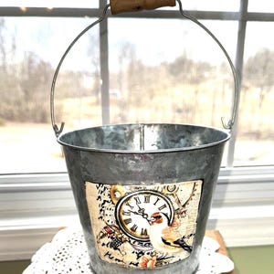 May include: A galvanized metal bucket with a metal handle and a wooden grip. The bucket features a decorative label with a clock, bird, and floral design. The bucket is sitting on a white lace doily.