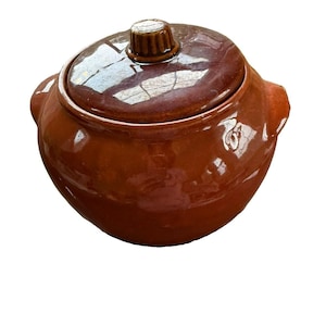 May include: A round, brown glazed ceramic pot with two side handles and a matching removable lid. The lid features a central knob with vertical ridges. The entire piece has a smooth, reflective finish and a consistent deep brown color throughout.