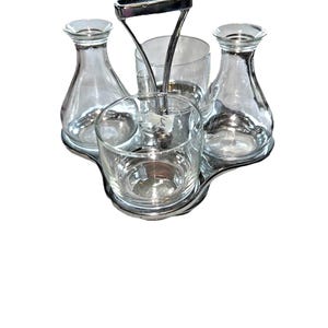 May include: A condiment server set featuring two clear glass carafes and two clear glass bowls arranged on a chrome-plated metal stand. The stand includes a central vertical handle for carrying, with a polished finish and a curved base.