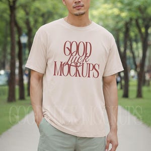 Könnte beinhalten: Beiges T-Shirt mit dem Text "GOOD Luck MOCKUPS" in burgunderfarbener Schrift. Das Model trägt hellgrüne Shorts und steht in einem Park mit Bäumen im Hintergrund.