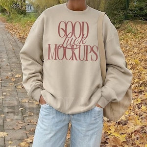 May include: A beige sweatshirt with the text "GOOD luck MOCKUPS" in burgundy lettering. The person is wearing light blue jeans and brown shoes. A beige shoulder bag is visible. The background features a walkway with fallen autumn leaves.