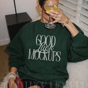 May include: A dark green sweatshirt with the text "GOOD luck MOCKUPS" in a white, elegant font. The sweatshirt is worn by a person holding a cocktail glass. The person is wearing jewelry and sitting on a white fur surface.