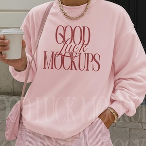 May include: A light pink sweatshirt with the text "GOOD Luck MOCKUPS" in a burgundy color. The person is wearing pink and white striped shorts and holding a white coffee cup. A gold chain necklace and bracelet are also visible.