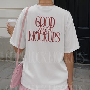 May include: A white t-shirt with the text "GOOD Luck MOCKUPS" in red script on the back. The person is wearing pink striped shorts and a pink shoulder bag. A white coffee cup is held in the left hand.