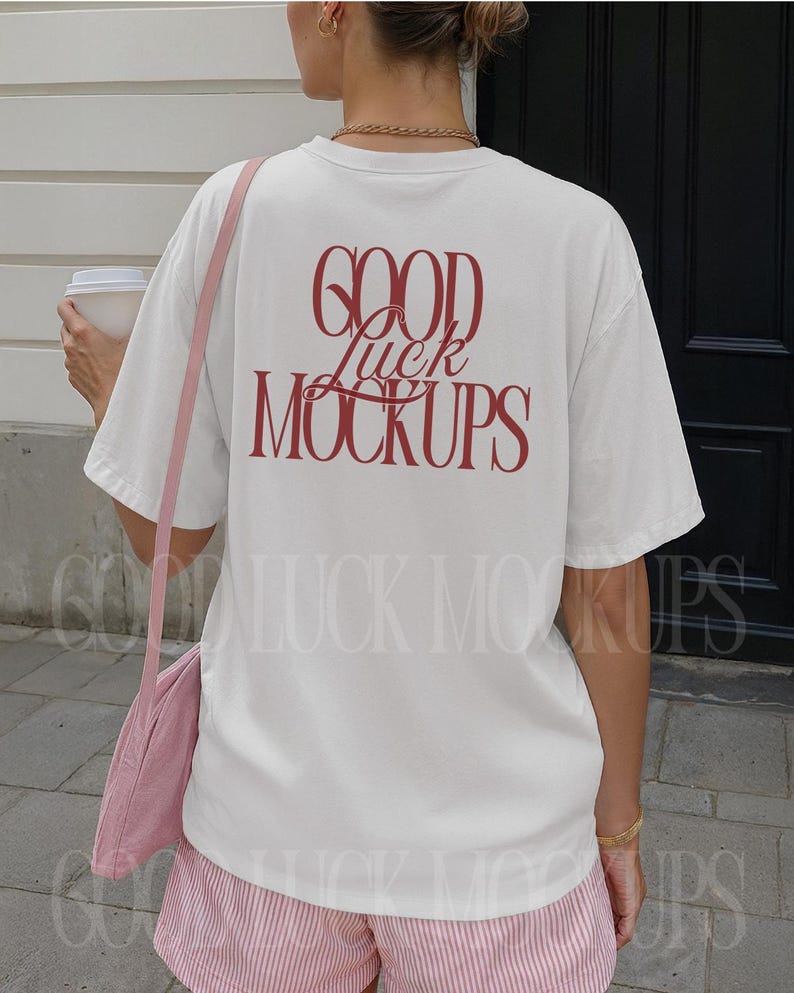 May include: A white t-shirt with the text "GOOD Luck MOCKUPS" in red script on the back. The person is wearing pink striped shorts and a pink shoulder bag. A white coffee cup is held in the left hand.
