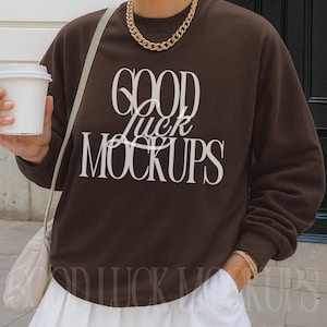 May include: A brown sweatshirt with the text "GOOD luck MOCKUPS" in white. The sweatshirt is paired with a white pleated skirt and gold jewelry. A white tote bag is slung over the shoulder.