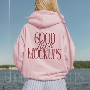 May include: A light pink hoodie with the text "GOOD Luck MOCKUPS" in a burgundy script. The model is wearing blue and white striped shorts. The background shows a body of water and a blue sky.