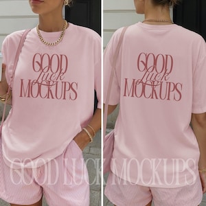 以下が含まれることがあります： 明るいピンク色のTシャツとショートパンツのセット。Tシャツには、前面と背面にバーガンディのスクリプトで「GOOD Luck MOCKUPS」の文字がプリントされています。ショートパンツはピンクと白のストライプです。モデルはゴールドのジュエリーとピンクのバッグを身に着けています。