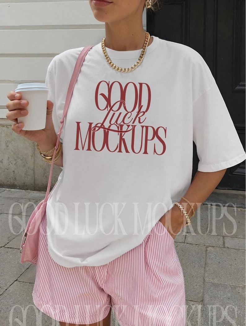 May include: White t-shirt with the text "GOOD LUCK MOCKUPS" in red, paired with pink and white striped shorts. The outfit is accessorized with gold jewelry and a pink purse.
