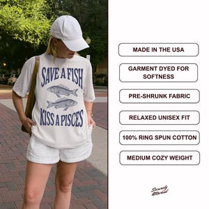May include: Cream-coloured t-shirt featuring the text "SAVE A FISH KISS A PISCES" and two fish illustrations in navy blue. The shirt is worn with white shorts and a white baseball cap. Made in the USA.