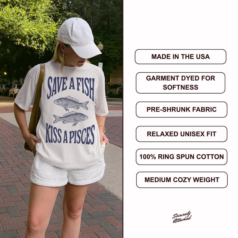May include: Cream-coloured t-shirt featuring the text "SAVE A FISH KISS A PISCES" and two fish illustrations in navy blue. The shirt is worn with white shorts and a white baseball cap. Made in the USA.