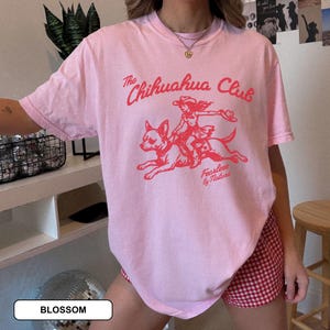 Puede incluir: Una camiseta de manga corta de color rosa claro con un estampado gr&aacute;fico rojo de una persona montando un chihuahua. El texto dice The Chihuahua Club y Fearless by Nature. La camiseta tiene cuello redondo y un corte holgado y relajado.