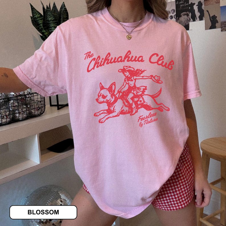 Puede incluir: Una camiseta de manga corta de color rosa claro con un estampado gr&aacute;fico rojo de una persona montando un chihuahua. El texto dice The Chihuahua Club y Fearless by Nature. La camiseta tiene cuello redondo y un corte holgado y relajado.