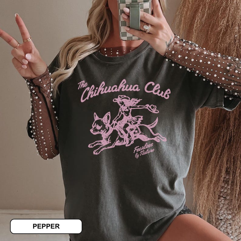 Puede incluir: Una camiseta gris carb&oacute;n con mangas largas transparentes adornadas con peque&ntilde;as perlas blancas. El gr&aacute;fico frontal muestra una ilustraci&oacute;n rosa de una persona montando un chihuahua con el texto The Chihuahua Club y Fearless by Nature.