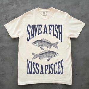 May include: Off-white t-shirt featuring the navy blue text "SAVE A FISH KISS A PISCES" and two fish illustrations. The tee has a crew neck and short sleeves, a casual style.