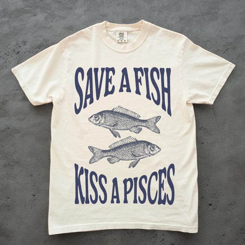 May include: Off-white t-shirt featuring the navy blue text "SAVE A FISH KISS A PISCES" and two fish illustrations. The tee has a crew neck and short sleeves, a casual style.