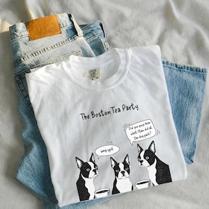 May include: A white t-shirt featuring a graphic of three Boston Terrier dogs with speech bubbles. The text reads The Boston Tea Party, omg spill, and Did you guys hear what Stan did at the dog park? The shirt is layered over a pair of light blue denim jeans.