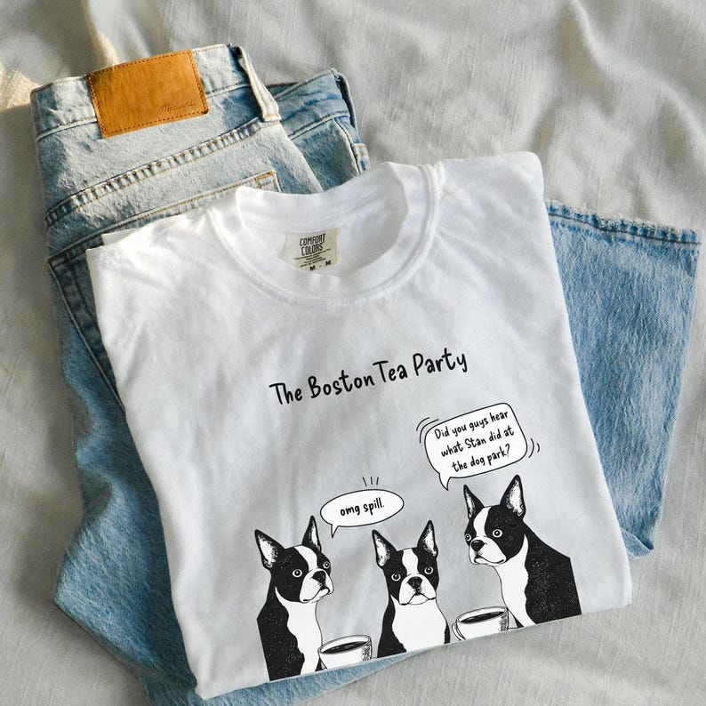 May include: A white t-shirt featuring a graphic of three Boston Terrier dogs with speech bubbles. The text reads The Boston Tea Party, omg spill, and Did you guys hear what Stan did at the dog park? The shirt is layered over a pair of light blue denim jeans.