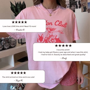 May include: A light pink t-shirt featuring a red graphic print of a person wearing a cowboy hat riding a dog, with the text "on Club" visible above the graphic. The shirt has a crew neckline and short sleeves, worn with red and white checkered shorts.
