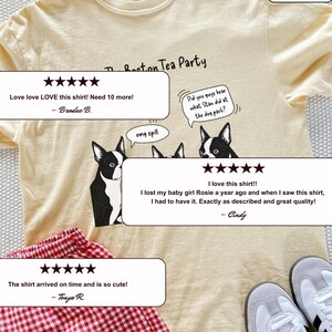 May include: A pale yellow t-shirt featuring a black and white graphic of three Boston Terrier dogs with speech bubbles that read The Boston Tea Party, omg spill, and Did you guys hear what Stan did at the dog park? The shirt is paired with red gingham shorts.