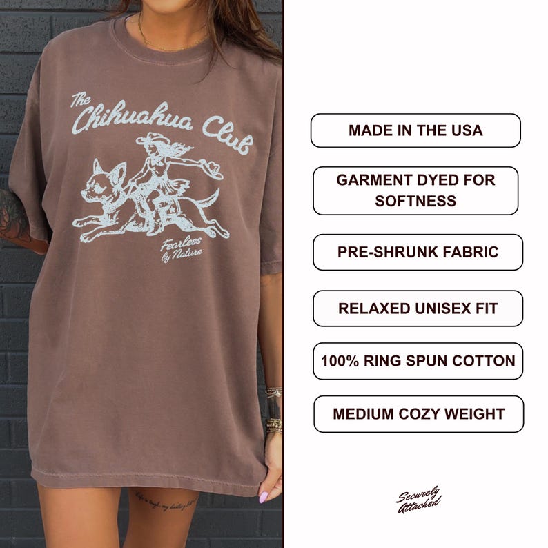 Puede incluir: Una camiseta marr&oacute;n con un gr&aacute;fico azul claro de una persona montando un chihuahua con el texto The Chihuahua Club y Fearless by Nature. Los cuadros de texto enumeran: Made in the USA, Garment Dyed for Softness, Pre-Shrunk Fabric, Relaxed Unisex Fit, 100% Ring Spun Cotton, Medium Cozy Weight.