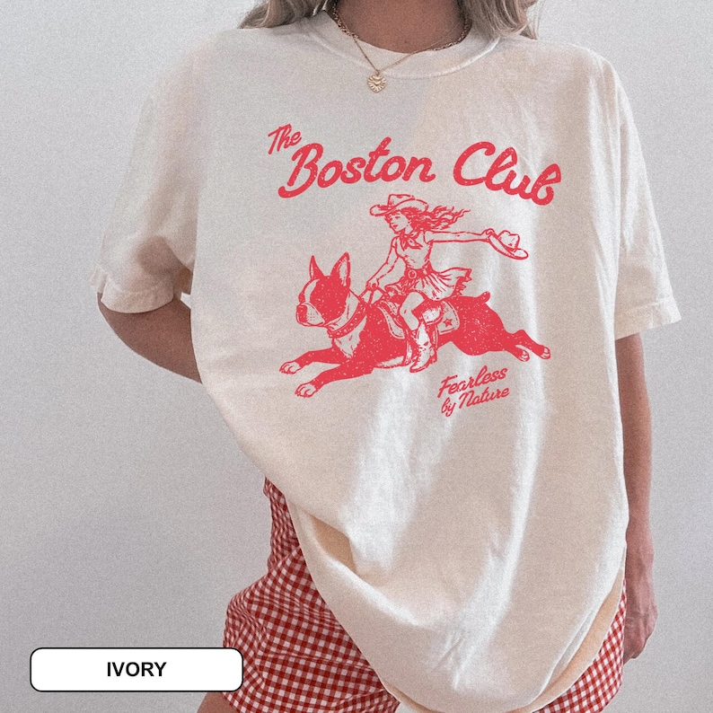 May include: An ivory short-sleeve t-shirt featuring a red graphic print of a person riding a Boston Terrier dog. The text reads The Boston Club and Fearless by Nature. The design has a distressed, vintage-inspired texture on the fabric.