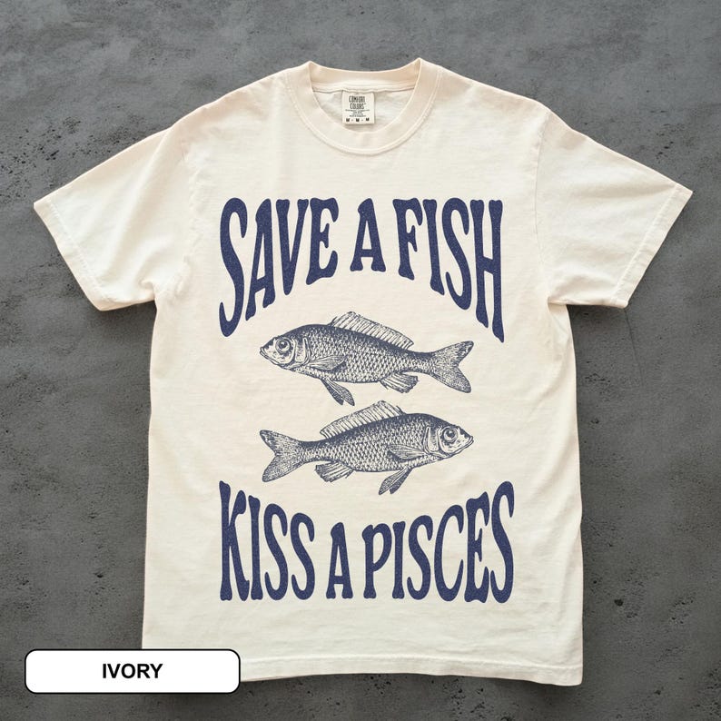 May include: Ivory t-shirt featuring the navy blue text "SAVE A FISH KISS A PISCES" and two fish illustrations. The tee has a crew neck and short sleeves. The shirt is made of a soft, comfortable material. The label at the bottom says "IVORY".