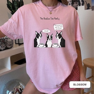 May include: A light pink t-shirt with a graphic of three Boston Terriers at a tea party. The dogs are black and white, with speech bubbles that say "omg spill" and "Did you guys hear what Stan did at the dog park?" The text "The Boston Tea Party" is above the graphic.