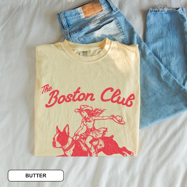 May include: A butter-colored t-shirt featuring a red graphic of a person in a cowboy hat riding a Boston Terrier. The text above the graphic reads The Boston Club in a cursive font. The shirt is paired with light blue denim jeans.