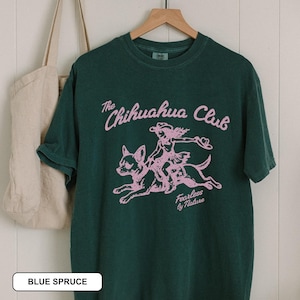 Puede incluir: Una camiseta de manga corta de color verde oscuro con un estampado gr&aacute;fico rosa claro de una persona montando un chihuahua. El texto dice The Chihuahua Club y Fearless by Nature. La camiseta est&aacute; en una percha de madera junto a un bolso de lona beige.