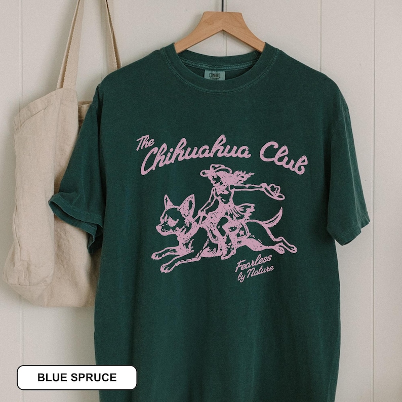 Puede incluir: Una camiseta de manga corta de color verde oscuro con un estampado gr&aacute;fico rosa claro de una persona montando un chihuahua. El texto dice The Chihuahua Club y Fearless by Nature. La camiseta est&aacute; en una percha de madera junto a un bolso de lona beige.