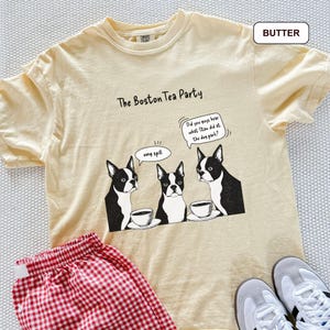 May include: A light yellow t-shirt with the text "The Boston Tea Party" and a graphic of three Boston Terriers with coffee cups. The shirt also has the text "Did you guys hear what Stan did at the dog park?" and "omg spill". Red and white checkered shorts are visible.