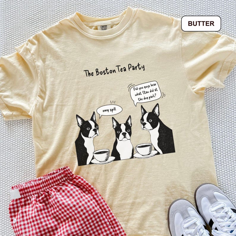 May include: A light yellow t-shirt with the text "The Boston Tea Party" and a graphic of three Boston Terriers with coffee cups. The shirt also has the text "Did you guys hear what Stan did at the dog park?" and "omg spill". Red and white checkered shorts are visible.