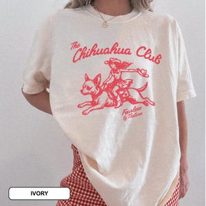 Puede incluir: Una camiseta de manga corta color marfil con un estampado gr&aacute;fico rojo de una persona montando un chihuahua. El texto dice The Chihuahua Club y Fearless by Nature. La camiseta tiene cuello redondo y un corte holgado y relajado.