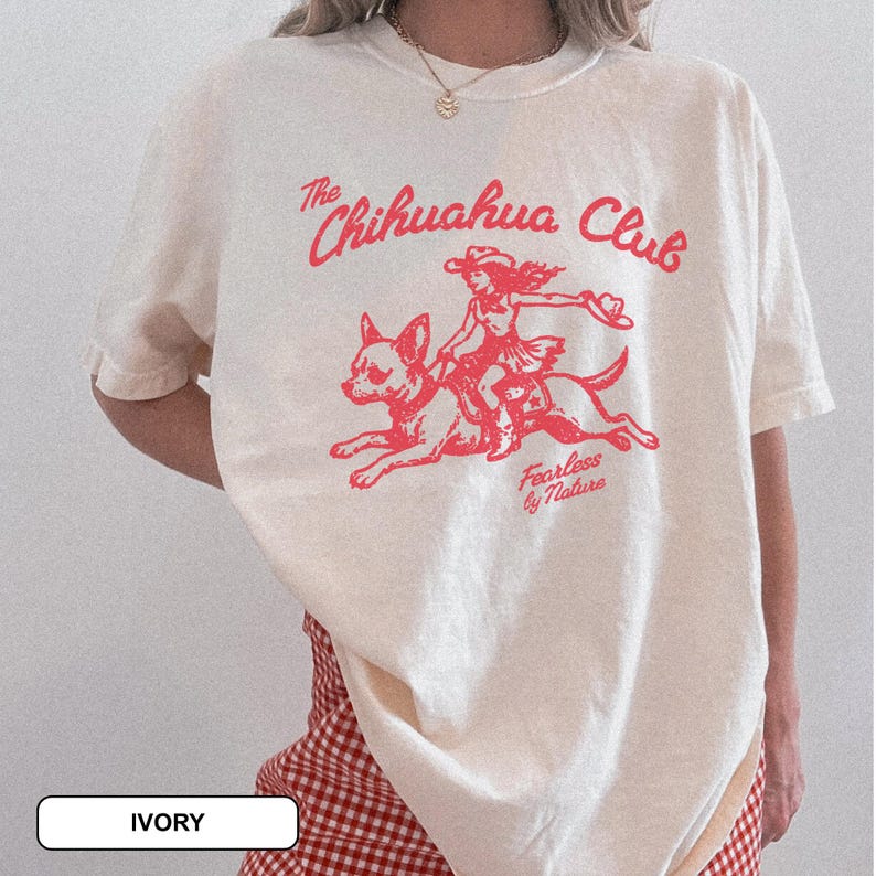 Puede incluir: Una camiseta de manga corta color marfil con un estampado gr&aacute;fico rojo de una persona montando un chihuahua. El texto dice The Chihuahua Club y Fearless by Nature. La camiseta tiene cuello redondo y un corte holgado y relajado.