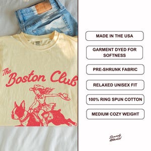 May include: A cream-colored t-shirt featuring red text that reads The Boston Club above a graphic of a person riding a Boston Terrier. The shirt is paired with blue denim jeans. Text boxes list features: Made in the USA, garment dyed, pre-shrunk, relaxed unisex fit, 100% ring spun cotton, and medium cozy weight.