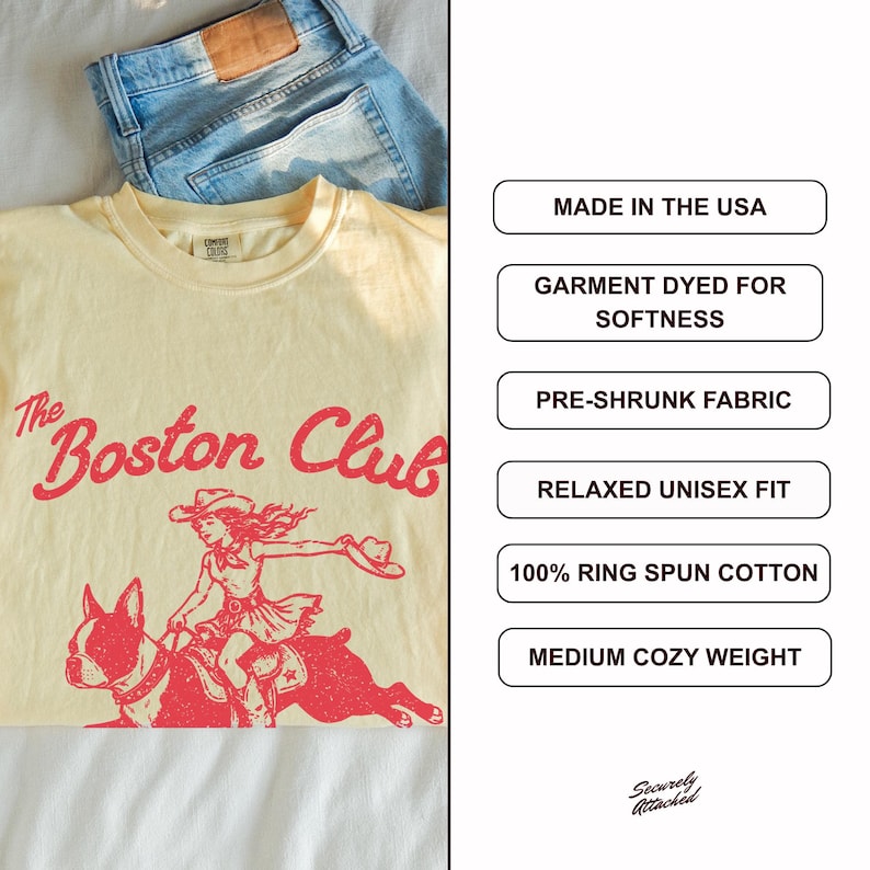 May include: A cream-colored t-shirt featuring red text that reads The Boston Club above a graphic of a person riding a Boston Terrier. The shirt is paired with blue denim jeans. Text boxes list features: Made in the USA, garment dyed, pre-shrunk, relaxed unisex fit, 100% ring spun cotton, and medium cozy weight.