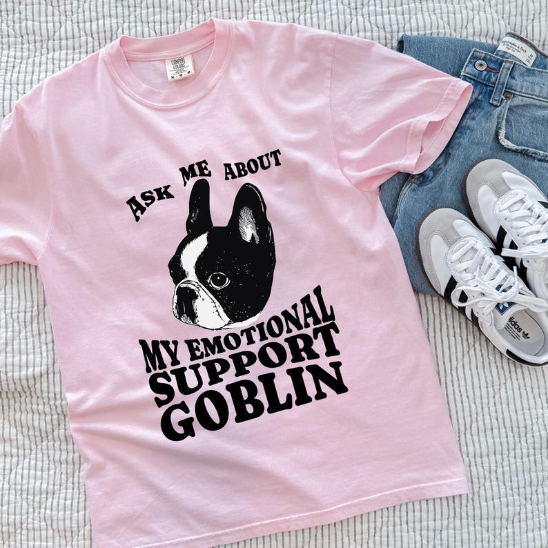 May include: A light pink t-shirt with the text "ASK ME ABOUT MY EMOTIONAL SUPPORT GOBLIN" and a black and white Boston Terrier graphic. The shirt is paired with blue jeans and white sneakers.