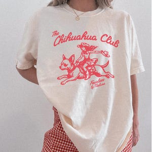 Puede incluir: Una camiseta de manga corta color blanco roto con un estampado gr&aacute;fico en rojo. El dise&ntilde;o muestra a una persona montando un chihuahua con el texto The Chihuahua Club y Fearless by Nature. La camiseta tiene cuello redondo y corte holgado.