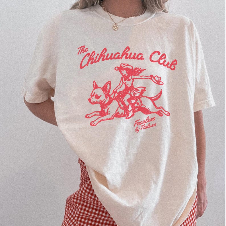 Puede incluir: Una camiseta de manga corta color blanco roto con un estampado gr&aacute;fico en rojo. El dise&ntilde;o muestra a una persona montando un chihuahua con el texto The Chihuahua Club y Fearless by Nature. La camiseta tiene cuello redondo y corte holgado.