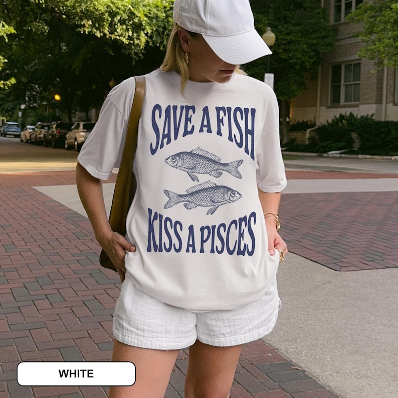 May include: A white t-shirt featuring the navy blue text "SAVE A FISH KISS A PISCES" and two fish illustrations. The shirt is worn with white shorts. The outfit has a relaxed, casual style.